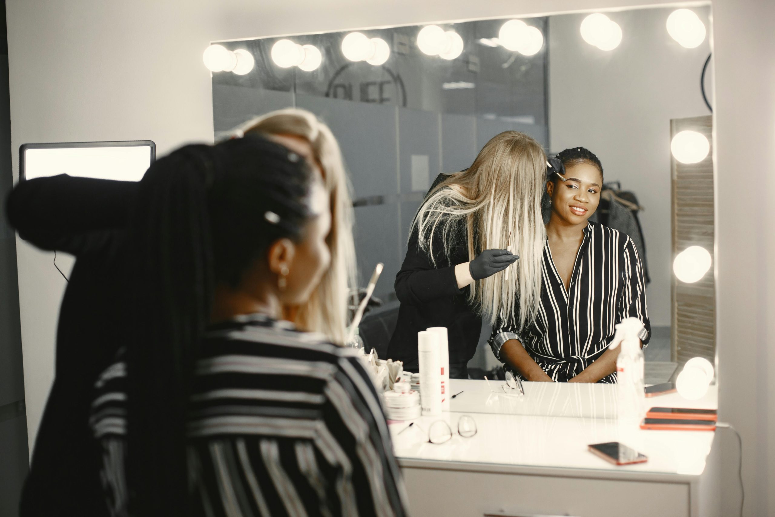 Makeup artist applies cosmetics to a smiling woman in a studio setting with bright vanity lights.